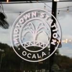 A white logo on a glass door has a fountain in the center with words in a circle around it reading, "Midtown Station Ocala."
