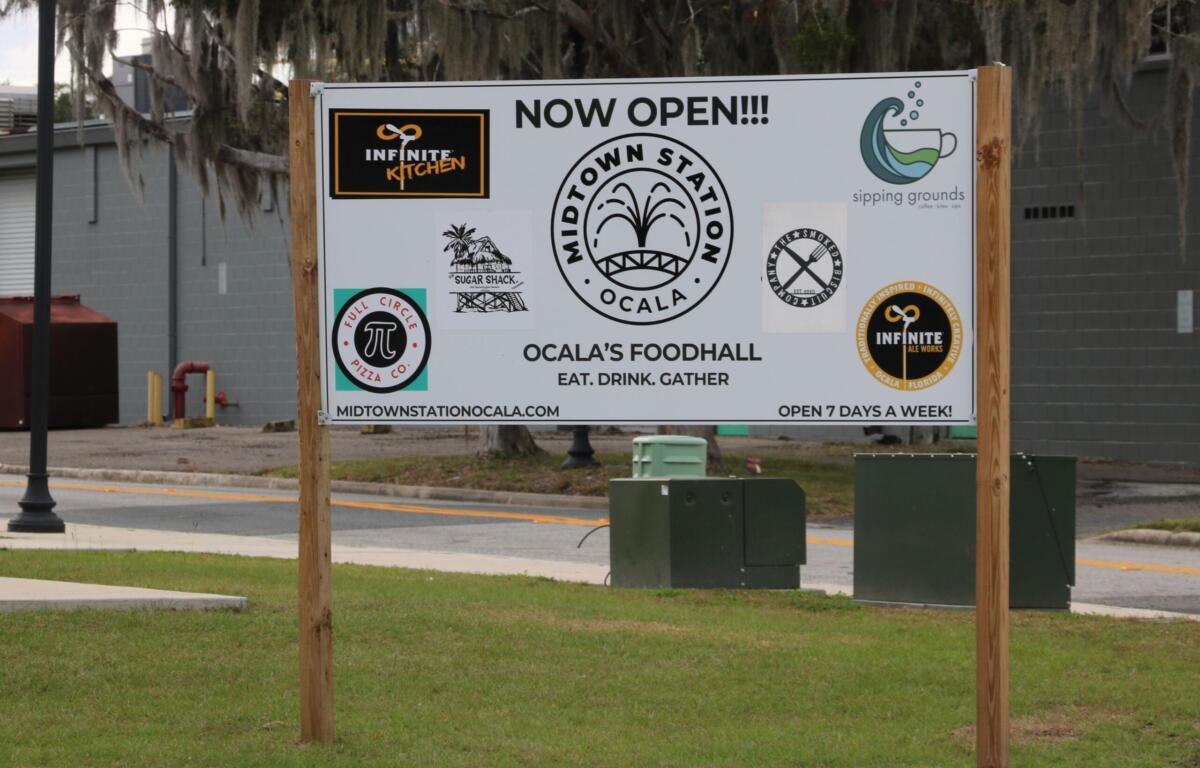 A white sign with multiple restaurant logos has "Now Open!!! Midtown Station Ocala. Ocala's Foodhall." In the background is a street view with a building behind it.