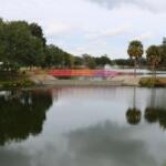 Lake Tuscawilla with trees and grass surrounding it, a bridge going over it and a fountain spraying upward behind it.