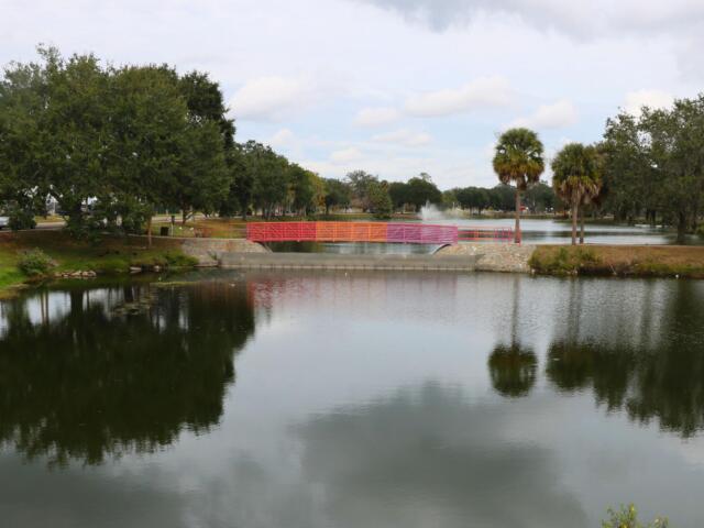 Lake Tuscawilla with trees and grass surrounding it, a bridge going over it and a fountain spraying upward behind it.