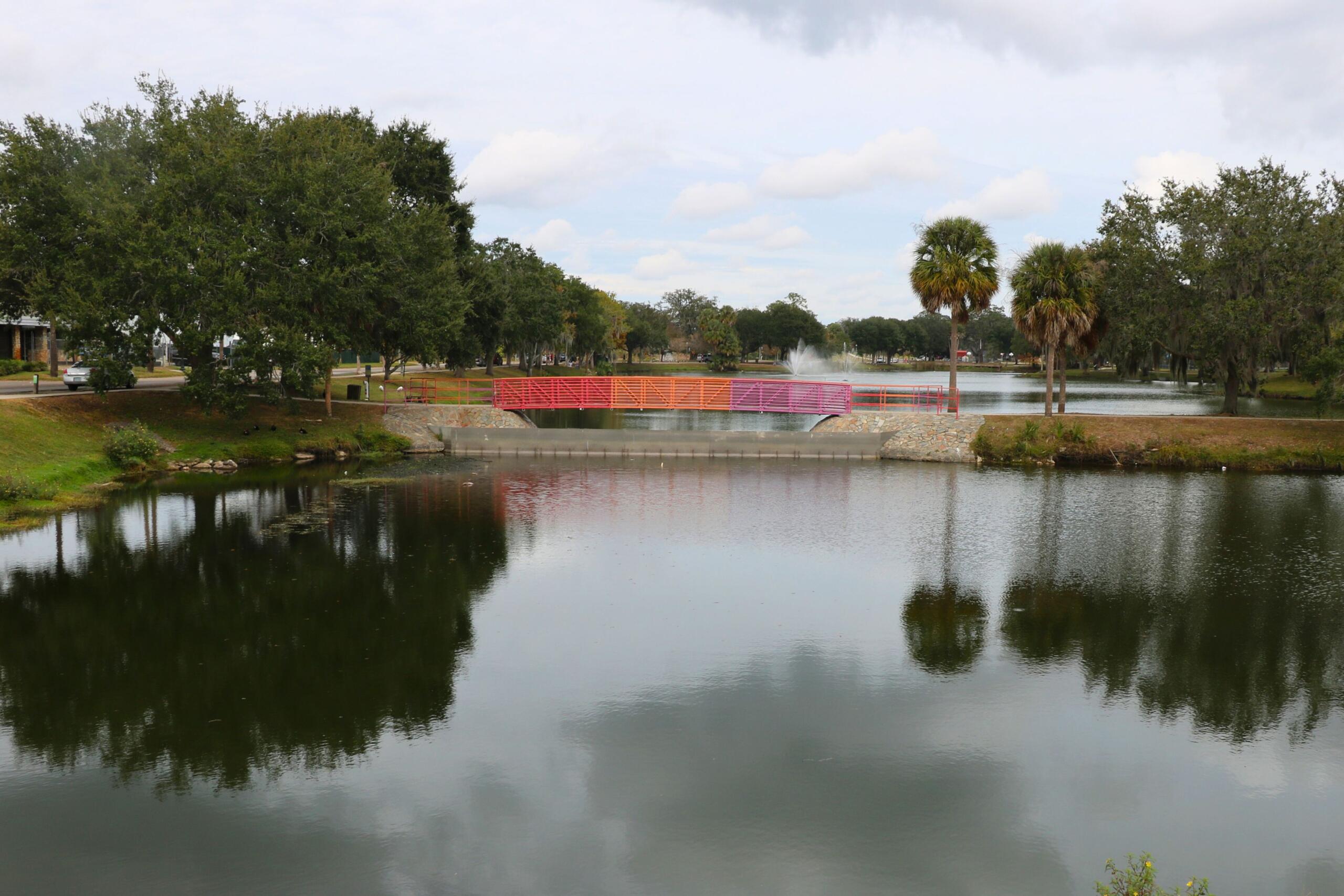 Lake Tuscawilla with trees and grass surrounding it, a bridge going over it and a fountain spraying upward behind it.