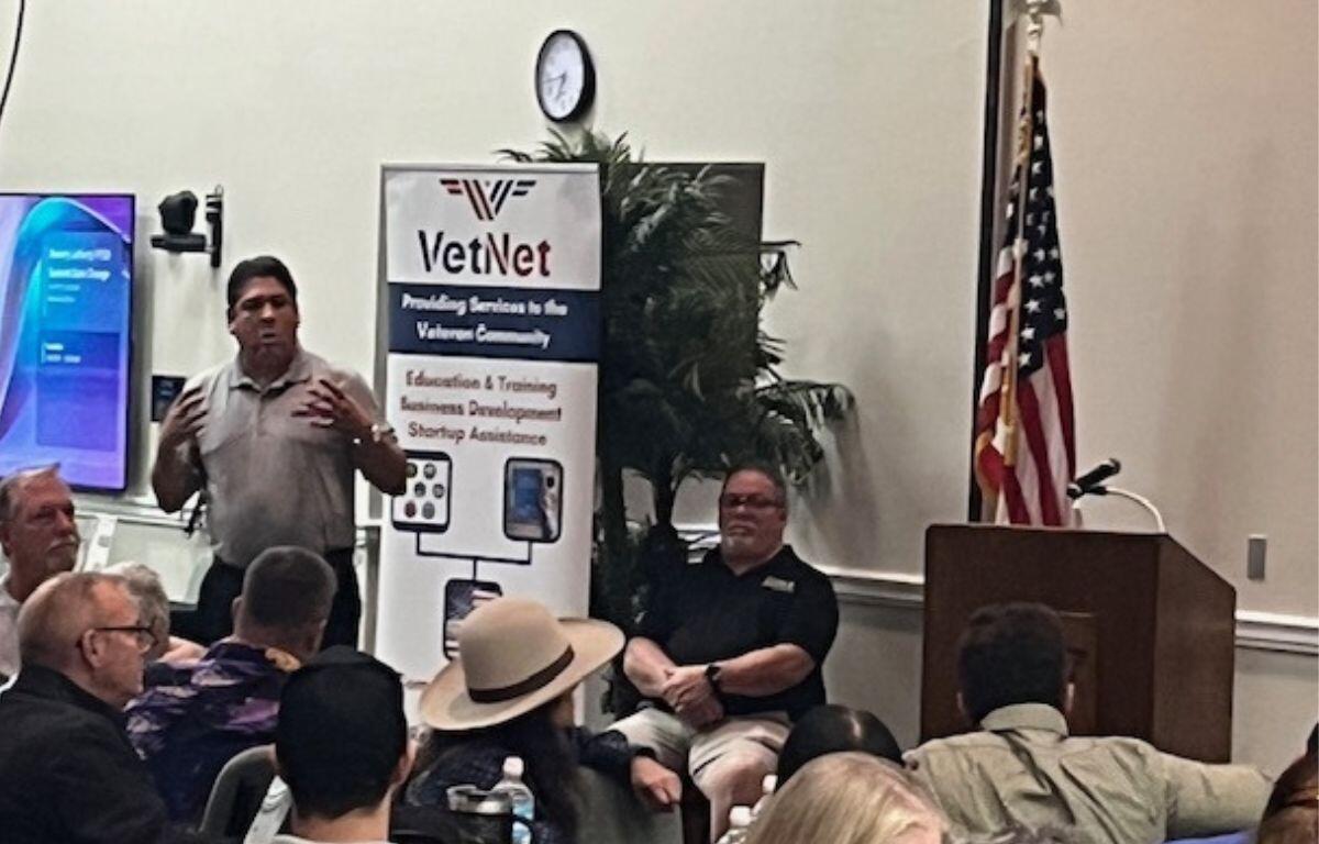 In the background, a placard with writing, a clock, a fake plant, a man sitting in a chair, and to the extreme right an American flag. The focal point a man in a gray polo, speaking and gesticulating.