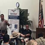 In the background, a placard with writing, a clock, a fake plant, a man sitting in a chair, and to the extreme right an American flag. The focal point a man in a gray polo, speaking and gesticulating.