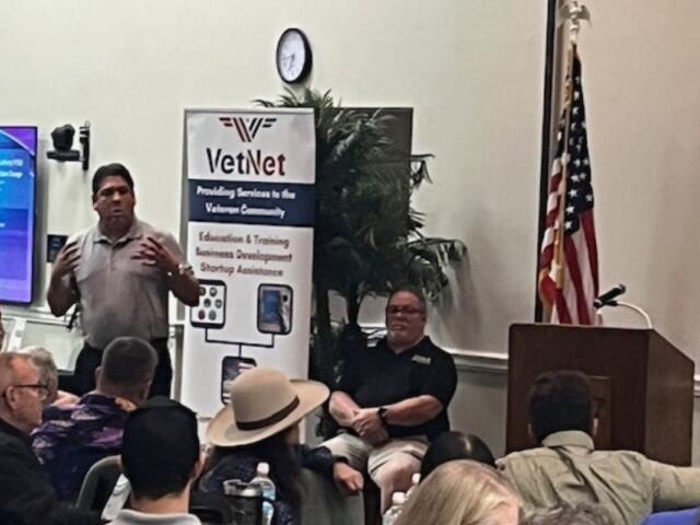 In the background, a placard with writing, a clock, a fake plant, a man sitting in a chair, and to the extreme right an American flag. The focal point a man in a gray polo, speaking and gesticulating.