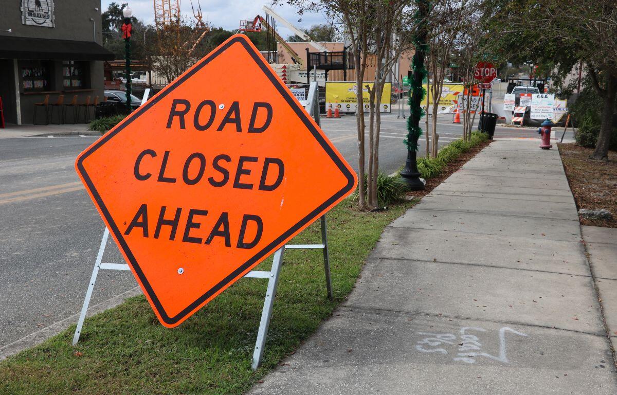 A large, orange, diamond-shaped sign with white supports reads, "Road Closed Ahead." To the right of it is a paved sidewalk leading to a construction site in the background and right has a road with a coffee shop on the corner of the road.