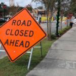 A large, orange, diamond-shaped sign with white supports reads, "Road Closed Ahead." To the right of it is a paved sidewalk leading to a construction site in the background and right has a road with a coffee shop on the corner of the road.