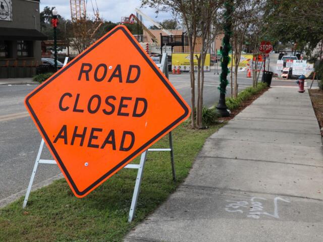 A large, orange, diamond-shaped sign with white supports reads, "Road Closed Ahead." To the right of it is a paved sidewalk leading to a construction site in the background and right has a road with a coffee shop on the corner of the road.