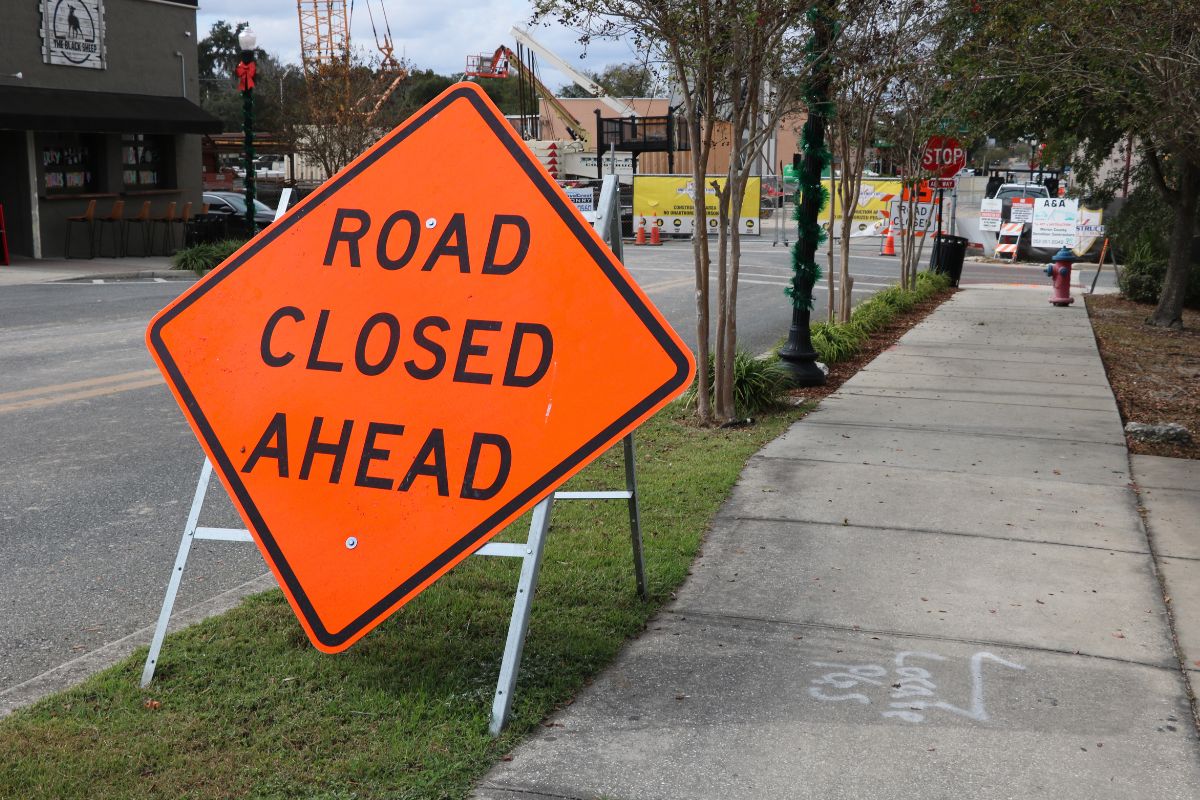 A large, orange, diamond-shaped sign with white supports reads, "Road Closed Ahead." To the right of it is a paved sidewalk leading to a construction site in the background and right has a road with a coffee shop on the corner of the road.