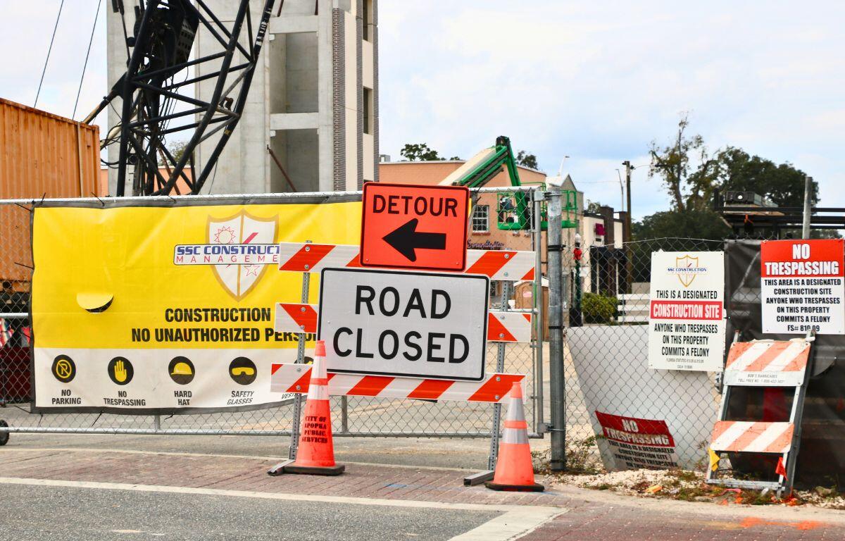 A construction site has multiple warning signs posted including an orange sign with a black arrow and black text reading, "Detour" and another white sign with text reading, "Road Closed."