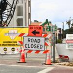 A construction site has multiple warning signs posted including an orange sign with a black arrow and black text reading, "Detour" and another white sign with text reading, "Road Closed."