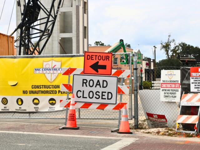 A construction site has multiple warning signs posted including an orange sign with a black arrow and black text reading, "Detour" and another white sign with text reading, "Road Closed."