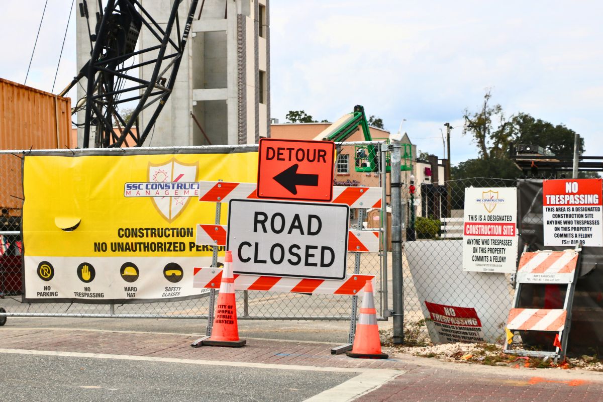 A construction site has multiple warning signs posted including an orange sign with a black arrow and black text reading, "Detour" and another white sign with text reading, "Road Closed."