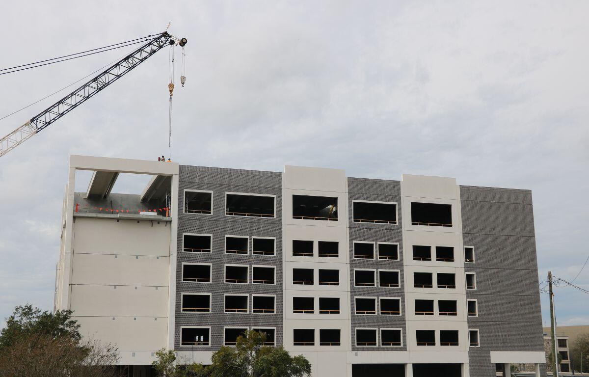 A beige and brick building is under construction with a crane hovering above it, providing materials.