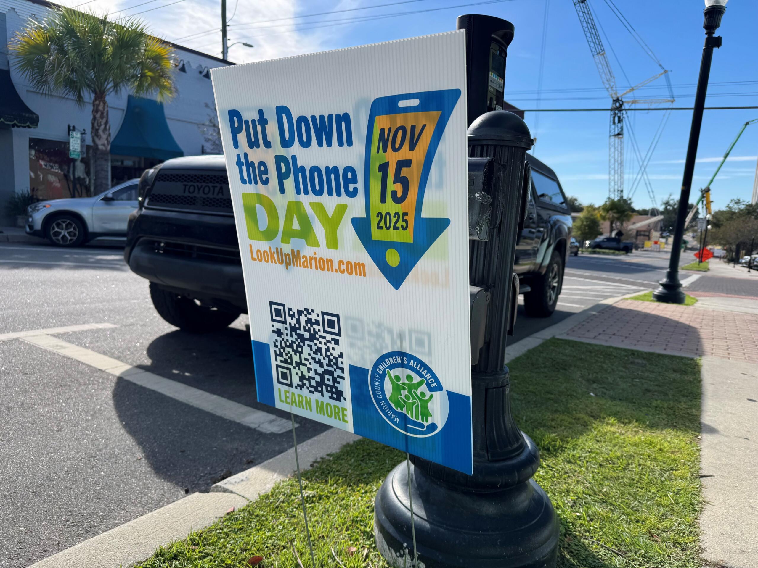 A "Put Down the Phone Day" yard sign placed in the grass in Downtown Ocala. The sign is in front of a parked car and parking meter.