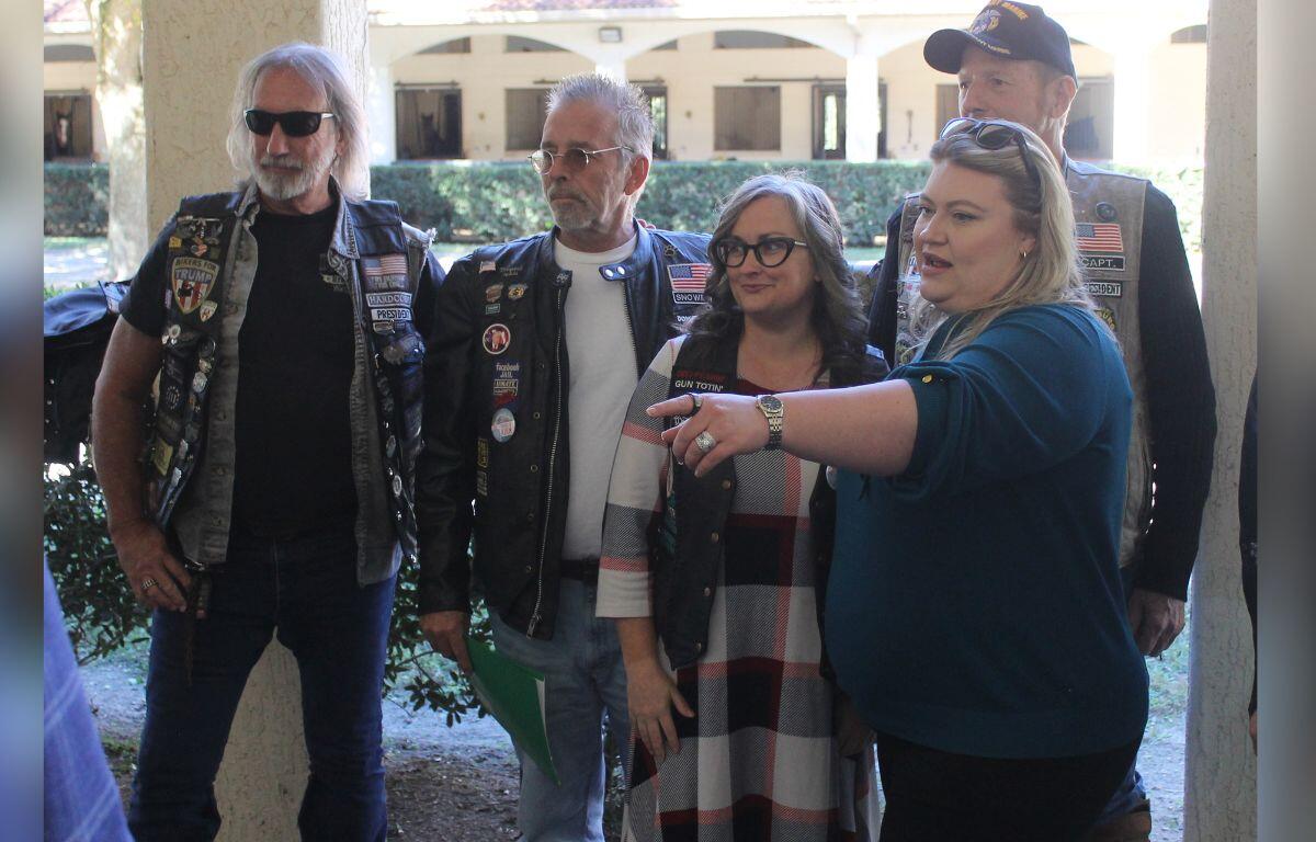 A group of people standing together, in front of an opening, with the woman on the far right extending her arm.