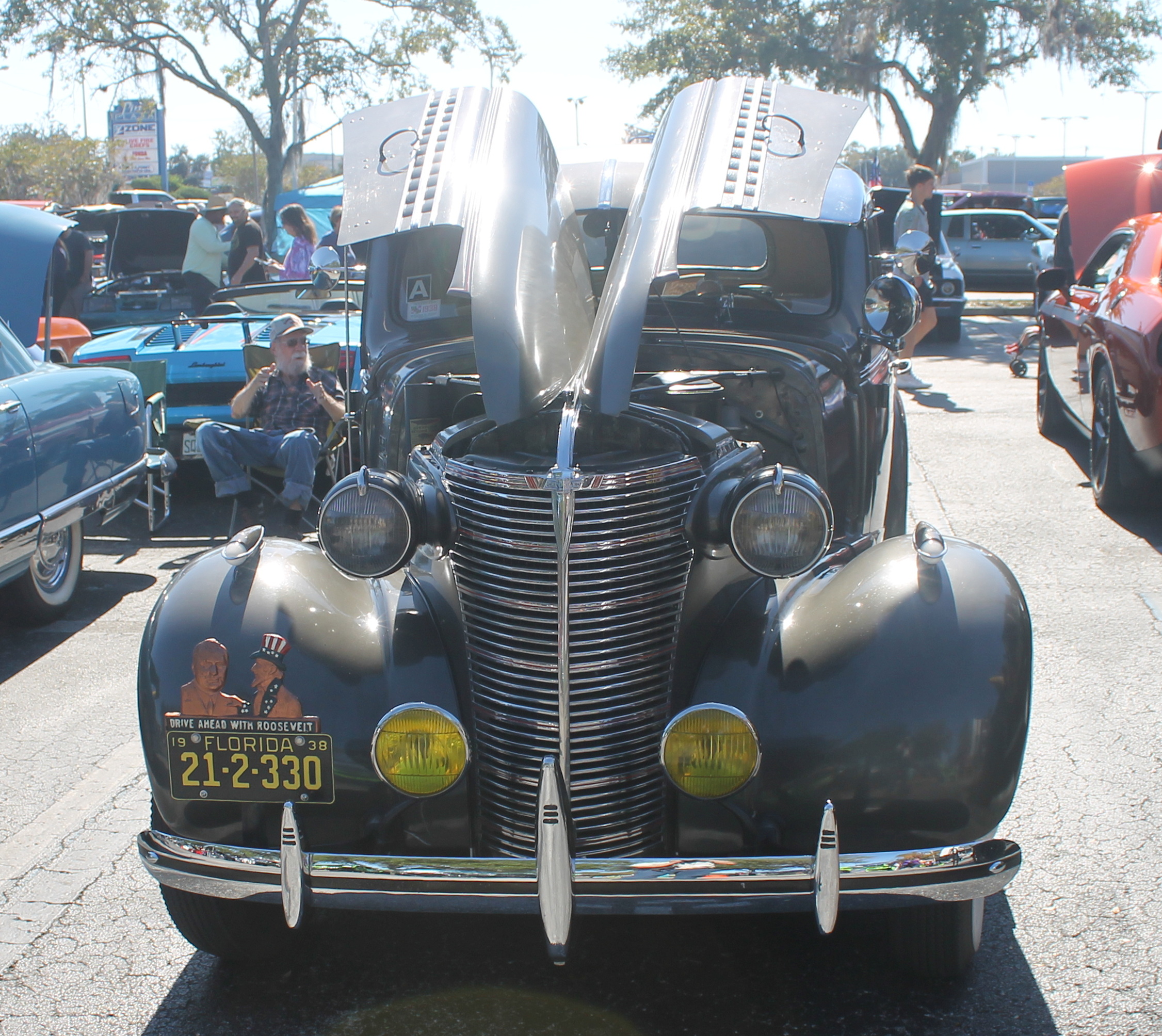 A black vehicle with its hood up, with license plate on the left side of the car, with a man sitting in the background on the left.