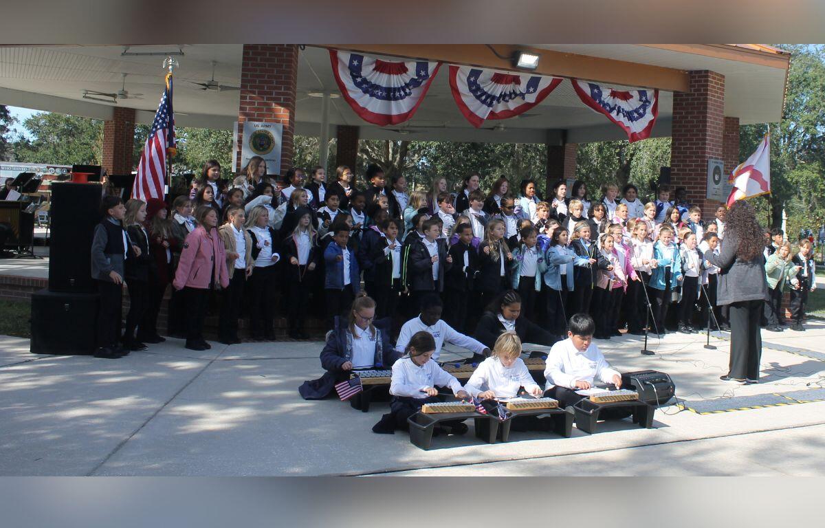 A group of people standing in front of a raised platform with an American flag to the left, banners above the people's heads, with students play vibraphones while sitting on a concrete walkway.