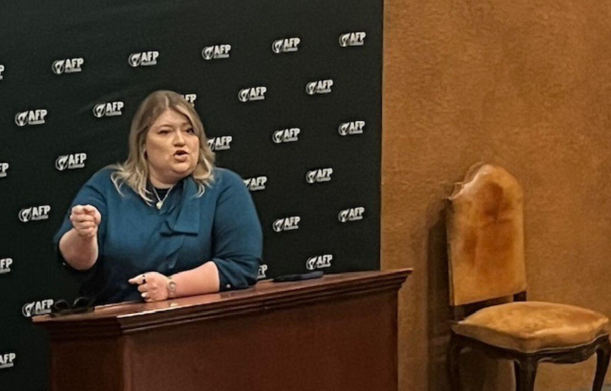 A woman behind a dais, with a banner with writing stretch across a wood panel wall, with an upholstered chair to the right.