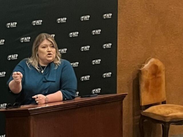 A woman behind a dais, with a banner with writing stretch across a wood panel wall, with an upholstered chair to the right.
