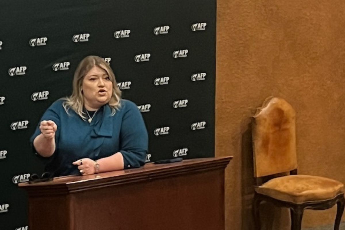 A woman behind a dais, with a banner with writing stretch across a wood panel wall, with an upholstered chair to the right.