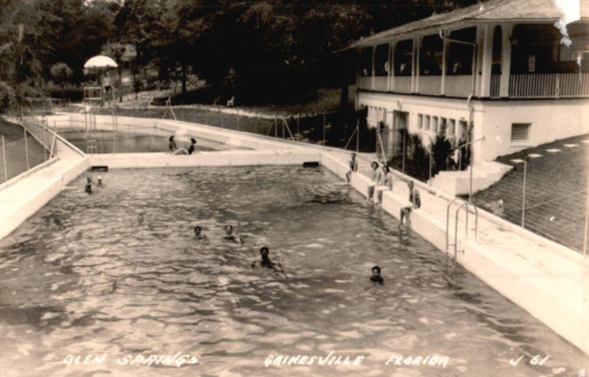 Historic image of swimmers enjoying the large spring-fed pool at Glen Springs, with the bathhouse stretching alongside the water and tree-covered banks rising behind it.
