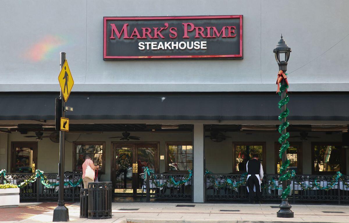 A restaurant has a fence surrounding it with Christmas decorations. A red, black and white sign above the restaurant entrance reads, "Mark's Prime Steakhouse."
