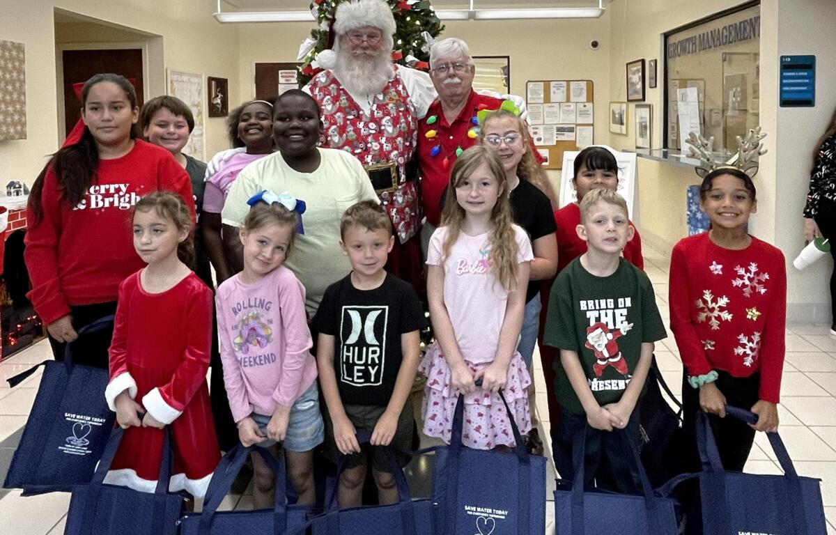 Villages Elementary of Lady Lake students have blue bags of gifts in hands. They pose with Santa with the Town Hall Christmas tree they decorated behind them.