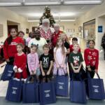Villages Elementary of Lady Lake students have blue bags of gifts in hands. They pose with Santa with the Town Hall Christmas tree they decorated behind them.