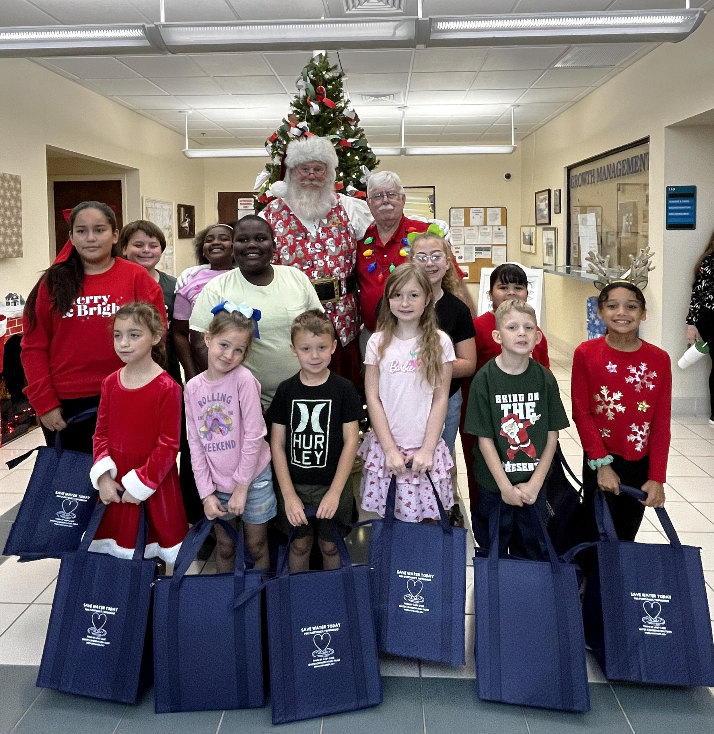 Villages Elementary of Lady Lake students have blue bags of gifts in hands. They pose with Santa with the Town Hall Christmas tree they decorated behind them.