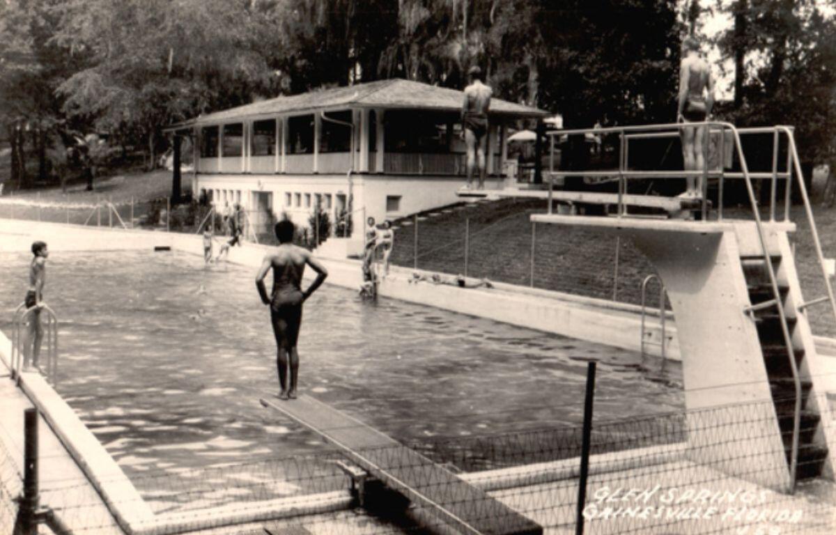 Historic photo of Glen Springs showing the pool deck, a lifeguard or staff member standing near the water, and the long, low bathhouse building in the background surrounded by trees.