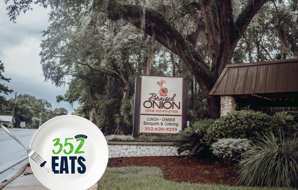The Braised Onion restaurant sign displayed along a landscaped walkway beneath large, shady trees.