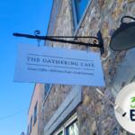 A white sign reading, "The Gathering Cafe" is hung on a historic stone building.