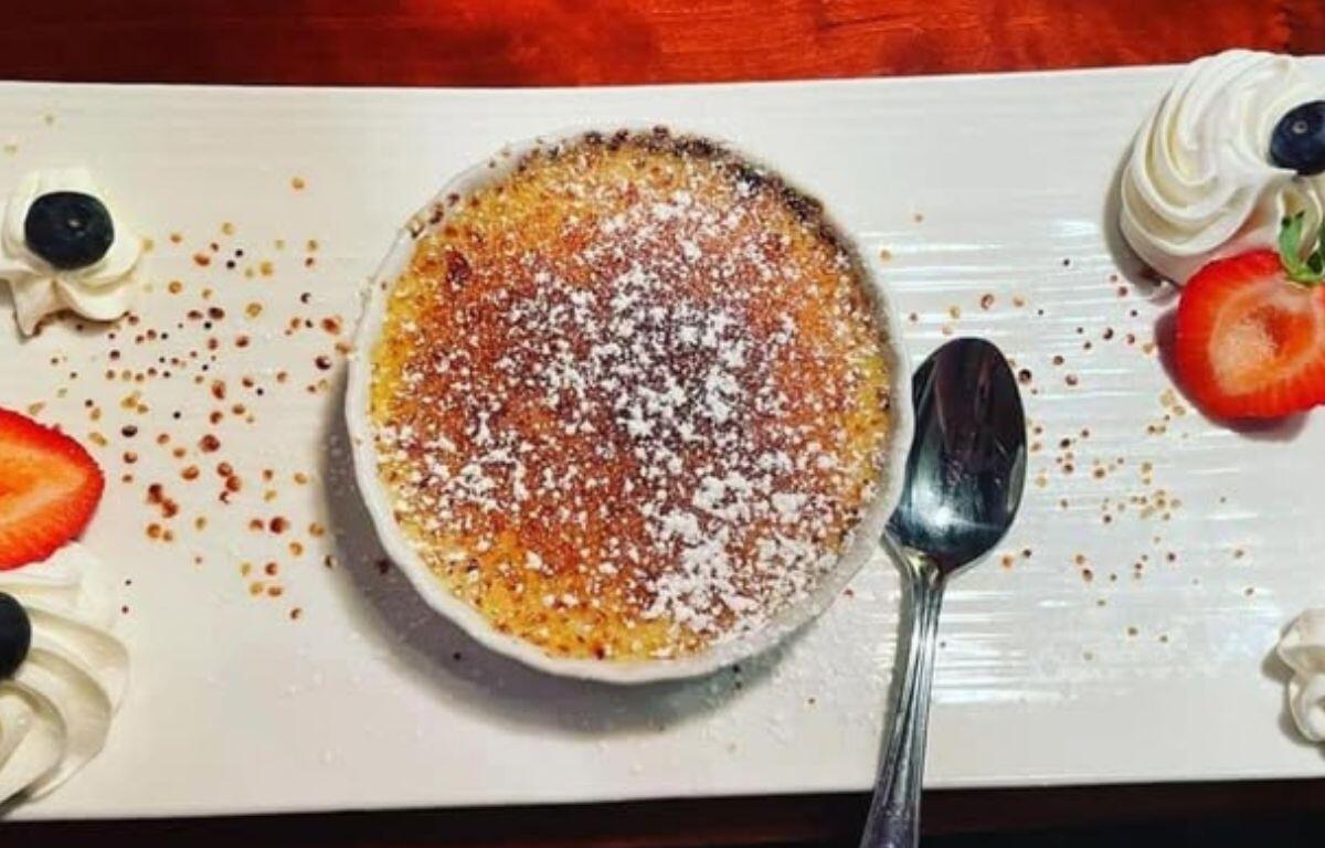 A close-up of a caramelized crème brûlée topped with powdered sugar, served in a white ramekin with a spoon beside it.
