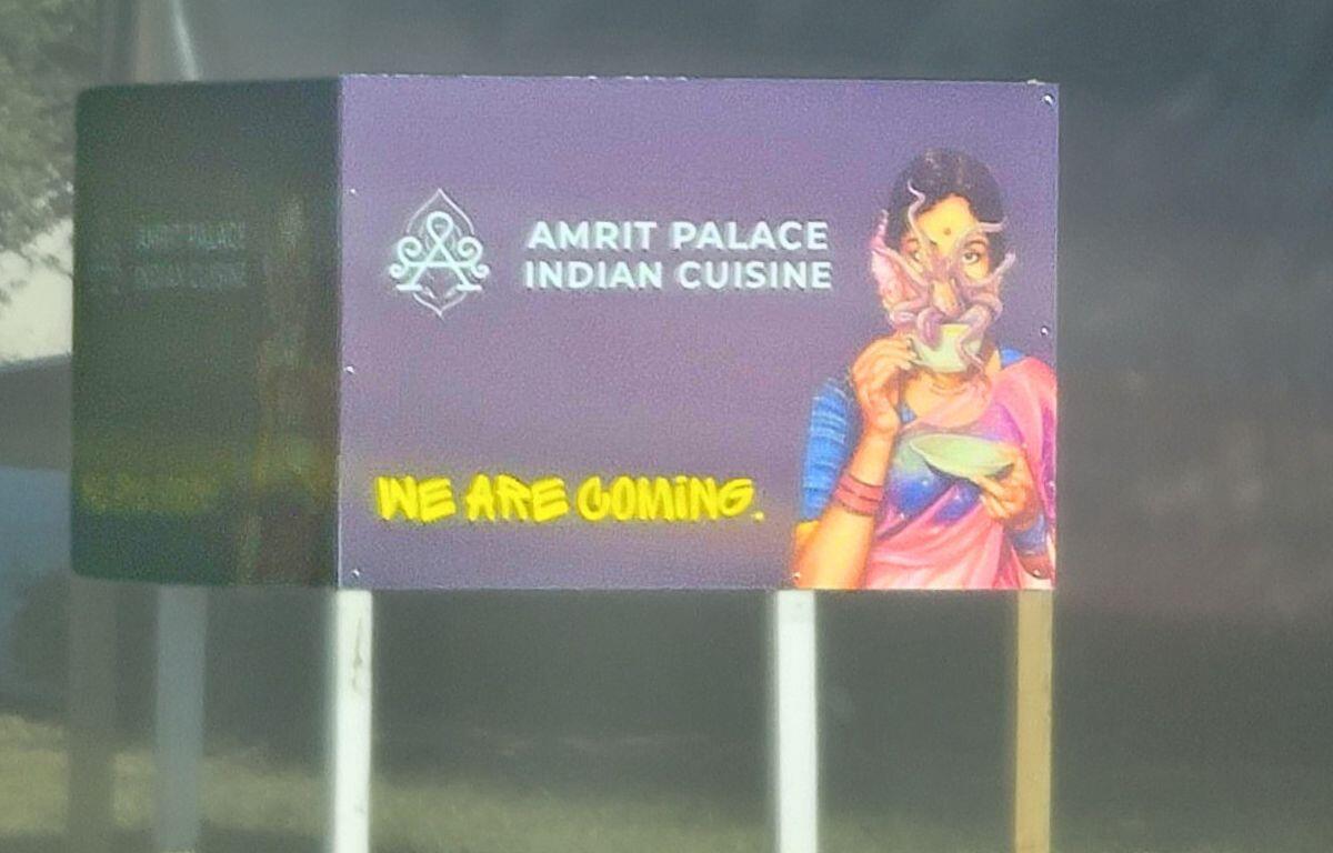A purple sign with white text reads, "Amrit Palace Indian Cuisine" with a drawing of a woman next to it. Yellow text on the sign reads, "We Are Coming." There is a wooded field in the background.