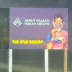 A purple sign with white text reads, "Amrit Palace Indian Cuisine" with a drawing of a woman next to it. Yellow text on the sign reads, "We Are Coming." There is a wooded field in the background.