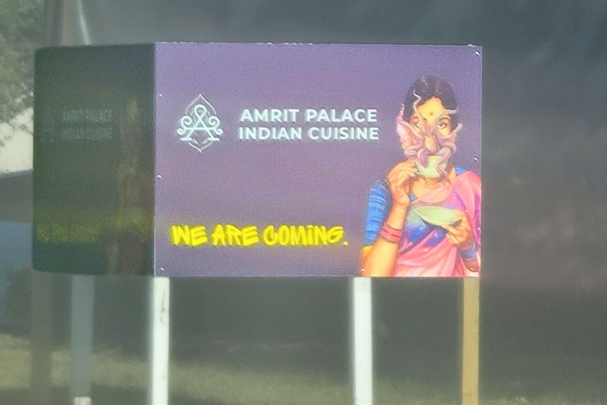 A purple sign with white text reads, "Amrit Palace Indian Cuisine" with a drawing of a woman next to it. Yellow text on the sign reads, "We Are Coming." There is a wooded field in the background.