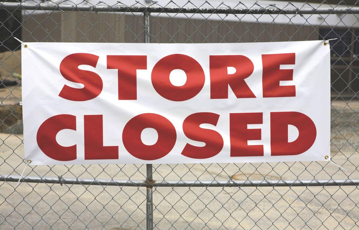 A building sits in the background while in the foreground, a metal fence blocks off the store with a banner hanging off of it, reading "Store Closed."