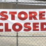 A building sits in the background while in the foreground, a metal fence blocks off the store with a banner hanging off of it, reading "Store Closed."
