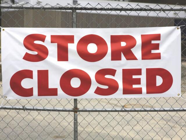 A building sits in the background while in the foreground, a metal fence blocks off the store with a banner hanging off of it, reading "Store Closed."