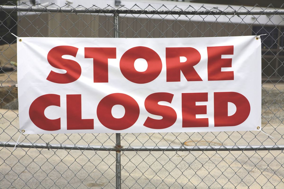 A building sits in the background while in the foreground, a metal fence blocks off the store with a banner hanging off of it, reading "Store Closed."
