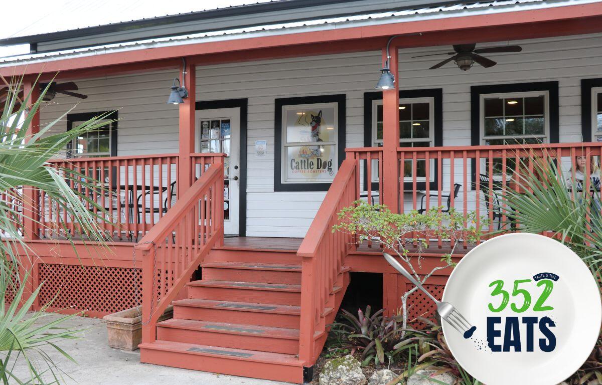 A historic house turned into a coffee shop features a sign in a window reading, "Cattle Dog Coffee Roasters." The sign has a graphic of a dog roaster a pot over a fire. A graphic in the foreground features a plate with a fork take a piece out of a blue letter. Green and blue letters read, "352EATS."