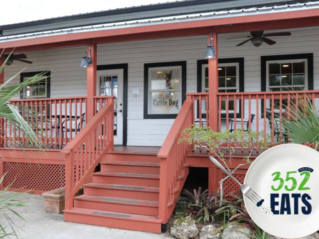 A historic house turned into a coffee shop features a sign in a window reading, "Cattle Dog Coffee Roasters." The sign has a graphic of a dog roaster a pot over a fire. A graphic in the foreground features a plate with a fork take a piece out of a blue letter. Green and blue letters read, "352EATS."