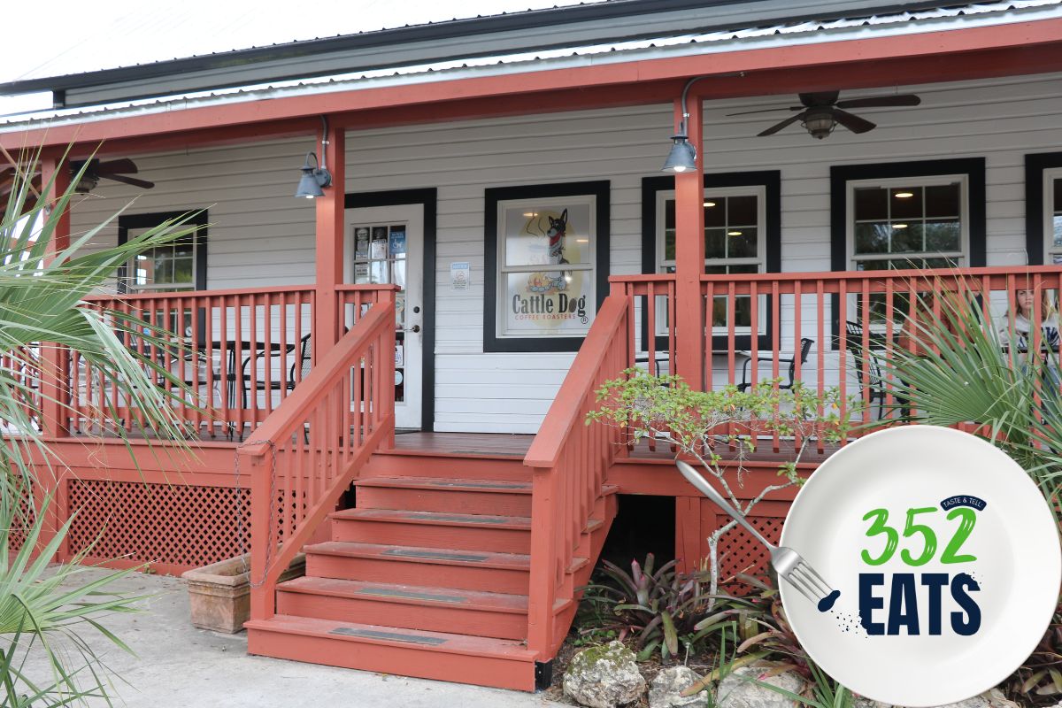 A historic house turned into a coffee shop features a sign in a window reading, "Cattle Dog Coffee Roasters." The sign has a graphic of a dog roaster a pot over a fire. A graphic in the foreground features a plate with a fork take a piece out of a blue letter. Green and blue letters read, "352EATS."