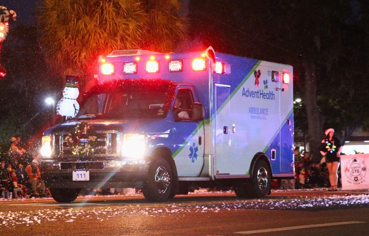 An ambulance with its lights on and a wreath in front of it drives down a dark road with fake snow.