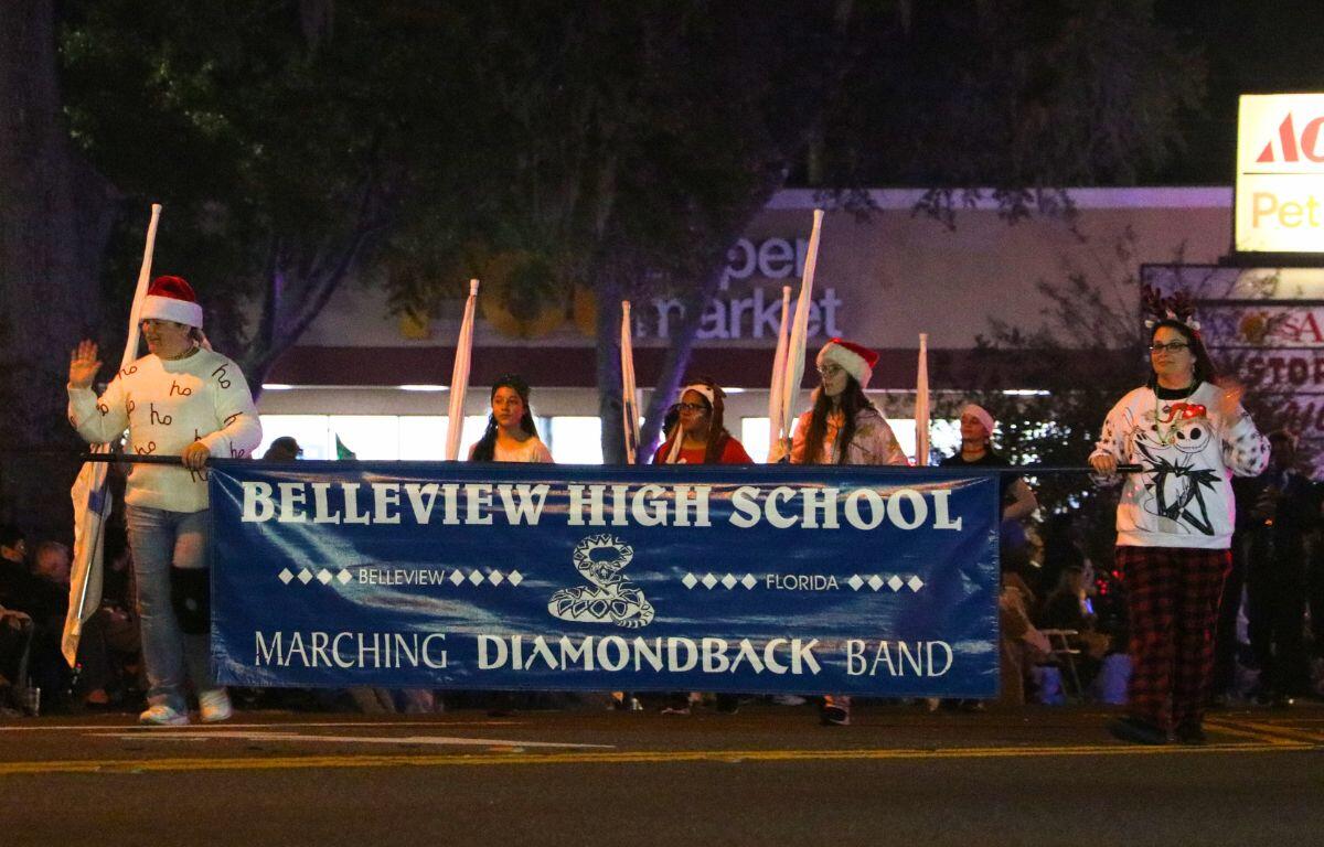 Students walk together with some twirling flags while the rest play instruments. Two students hold a blue banner reading, "Belleview High School."