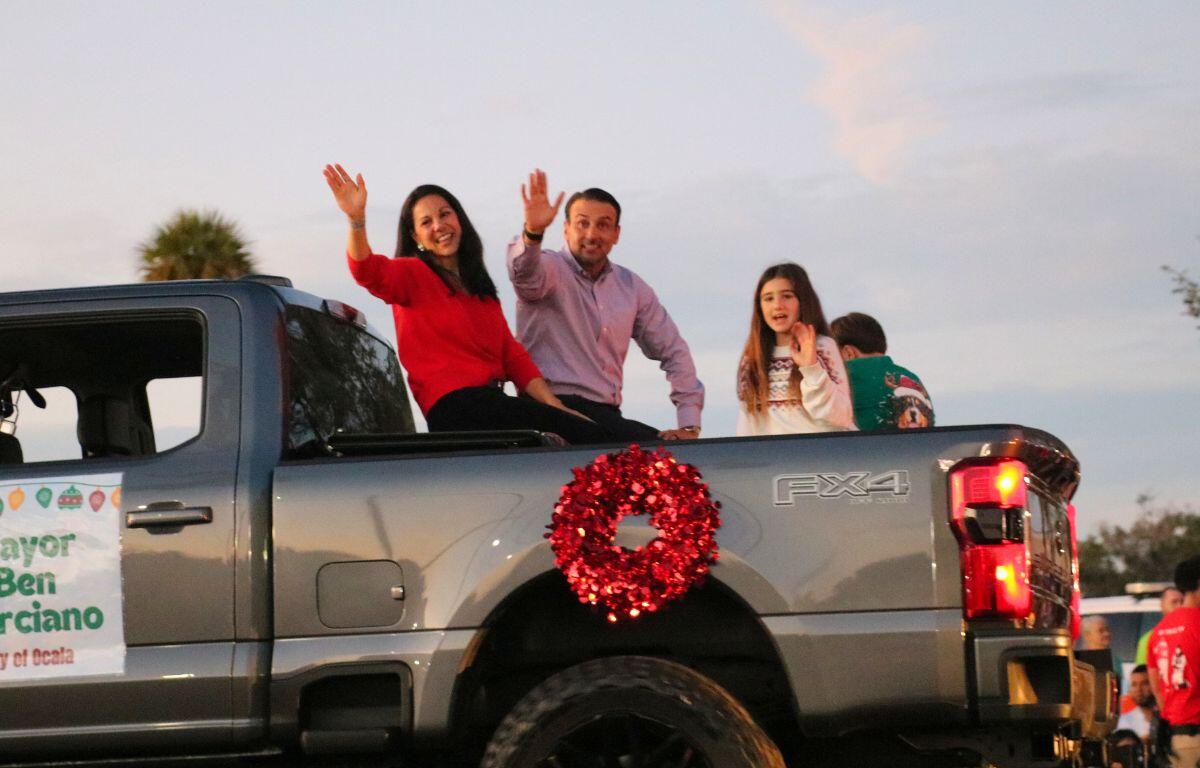 A family sits in the bed of a truck with a red wreath on it driving down a road.