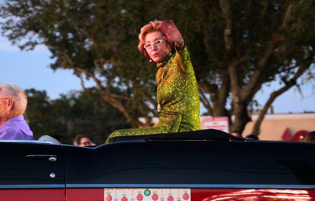 A woman in a green dress waves at a crowd of people while driving down a road.