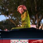 A woman in a green dress waves at a crowd of people while driving down a road.