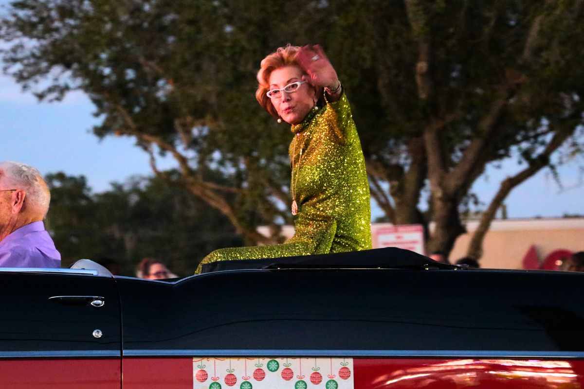 A woman in a green dress waves at a crowd of people while driving down a road.