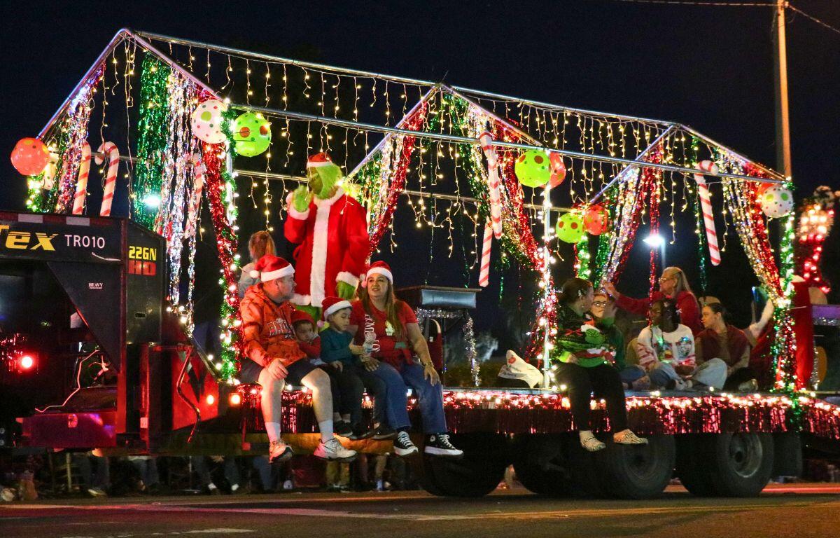 A truck features lights, candy canes and more Christmas decorations with people sitting on the edge as it drives down a dimly lit road.